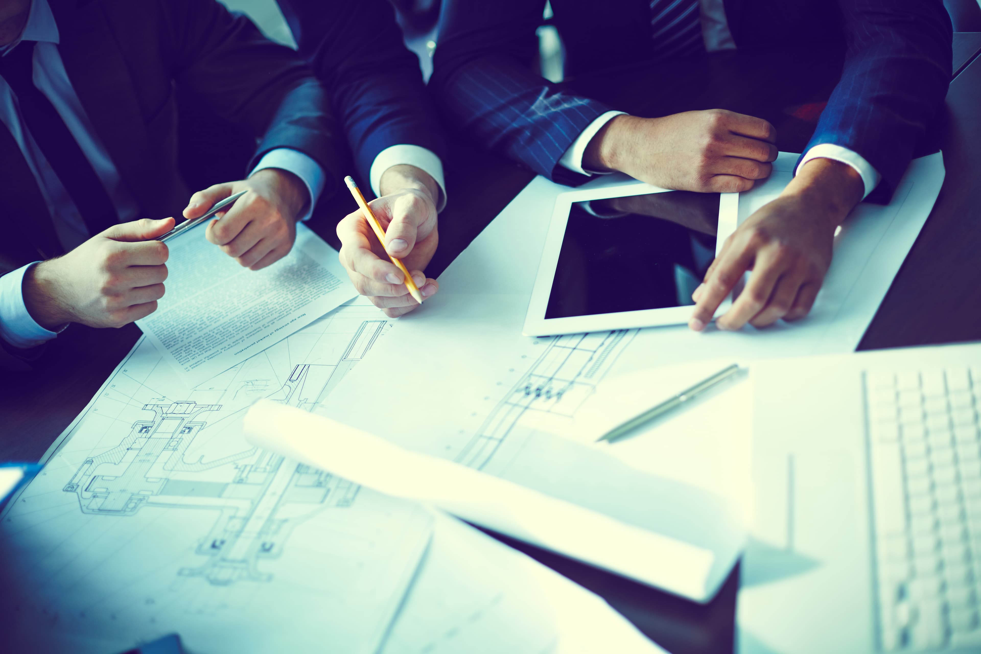 A group of people in an office, examining plans and notes on a table, using a tablet and documents, suggesting collaboration on an engineering or architectural project.