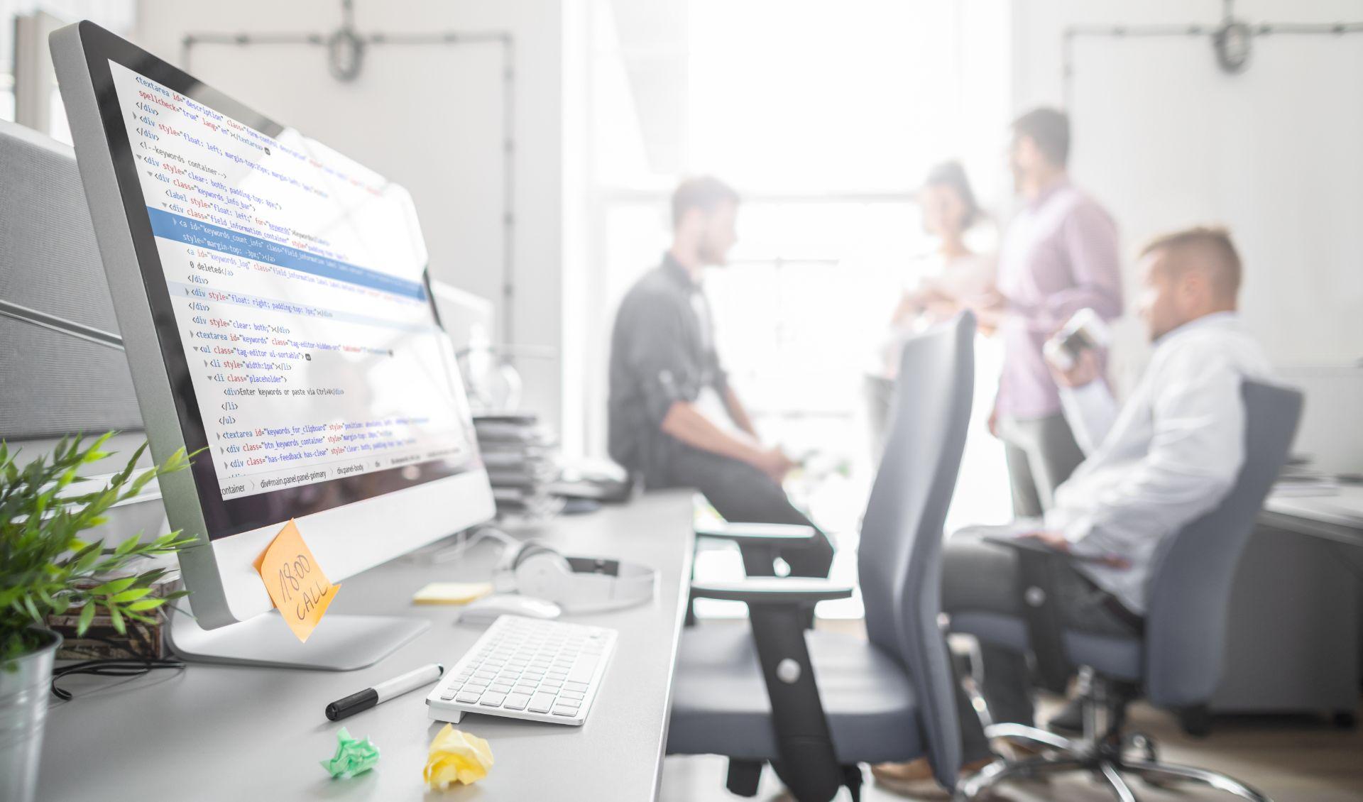 In the foreground is a computer with an application running. In the background, a group of people can be seen, looking like a team in an office socializing with each other.