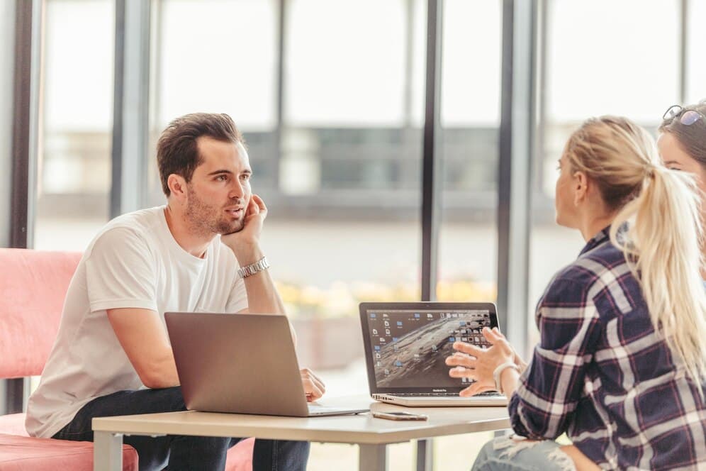 A group of young people in a modern office, talking at a table with laptops, suggesting a meeting or collaboration on a project.